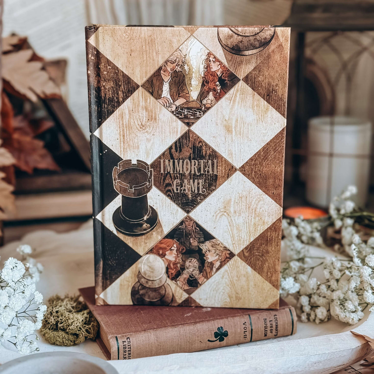 The hardcover book titled Immortal Game stands upright on top of an old book, surrounded by dried flowers and rustic decor. The cover is styled after a vintage brown and cream wooden chess board with small illustrated scenes and chess pieces.