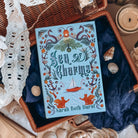 The front cover for the book Sea of Charms by Sarah Beth Durst shown resting on an open wicker basket with dark blue fabric and sea shells. The cover illustration shows the title surrounded by a variety of nautical iconography, like sea snakes, clamshells and pearls, anchors, and seaweed on a light blue background.