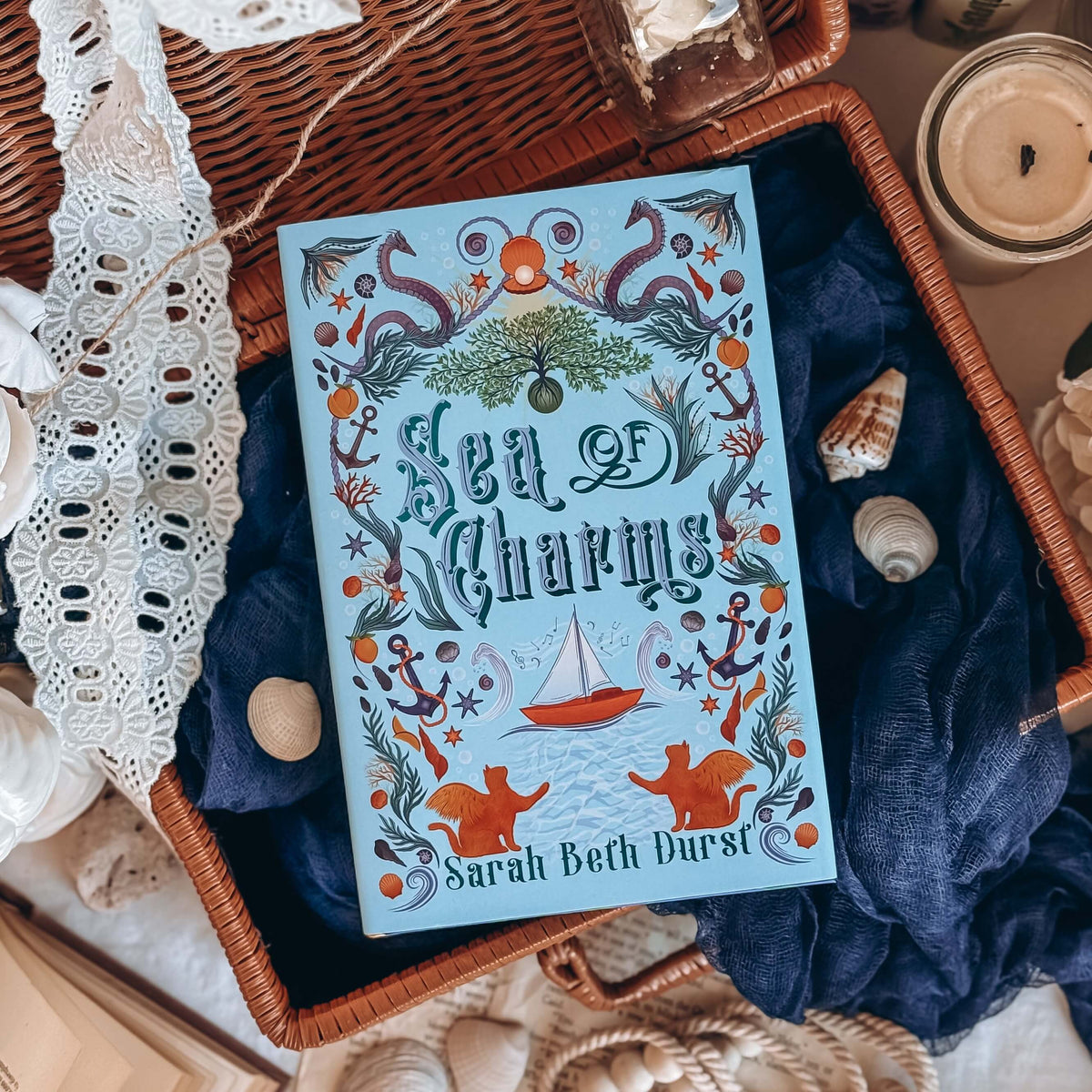The front cover for the book Sea of Charms by Sarah Beth Durst shown resting on an open wicker basket with dark blue fabric and sea shells. The cover illustration shows the title surrounded by a variety of nautical iconography, like sea snakes, clamshells and pearls, anchors, and seaweed on a light blue background.