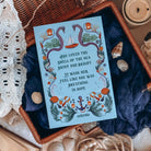 The back cover for the book Sea of Charms by Sarah Beth Durst shown resting on an open wicker basket with dark blue fabric and sea shells. The back illustration shows a quote surrounded by a variety of nautical iconography, like sea snakes, clamshells and pearls, anchors, and seaweed on a light blue background.