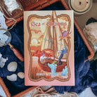 The front reversible dust jacket for the book Sea of Charms by Sarah Beth Durst shown resting on an open wicker basket with a dark blue fabric and sea shells. The illustration shows two characters on a small sailboat with a blue sea creature floating by. 
