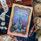 The back reversible dust jacket for the book Sea of Charms by Sarah Beth Durst shown resting on an open wicker basket with a dark blue fabric and sea shells. Back jacket shows an illustated sailboat scene at night with a sea creature floating by. The quote 'At some point, you just had to hoist the sail and go' is shown in the night sky.