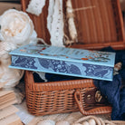 The stenciled edges for the book Sea of Charms by Sarah Beth Durst styled in an open wicker basket surrounded by decorative items. The nautical-inspired illustrated edges depict kelp and seashells on a dark blue background.  