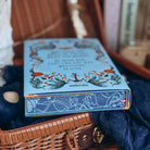 A close-up of the stenciled edges for the book Sea of Charms by Sarah Beth Durst styled in an open wicker basket surrounded by decorative items. The nautical-inspired illustrated edges depict kelp and seashells on a dark blue background.  