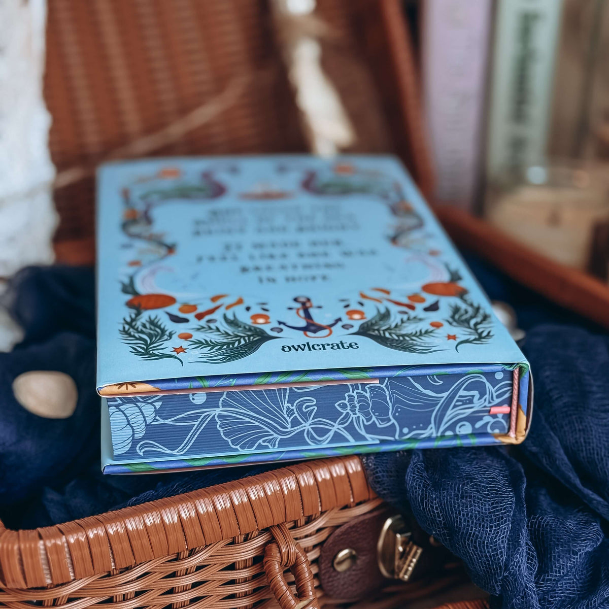 A close-up of the stenciled edges for the book Sea of Charms by Sarah Beth Durst styled in an open wicker basket surrounded by decorative items. The nautical-inspired illustrated edges depict kelp and seashells on a dark blue background.  