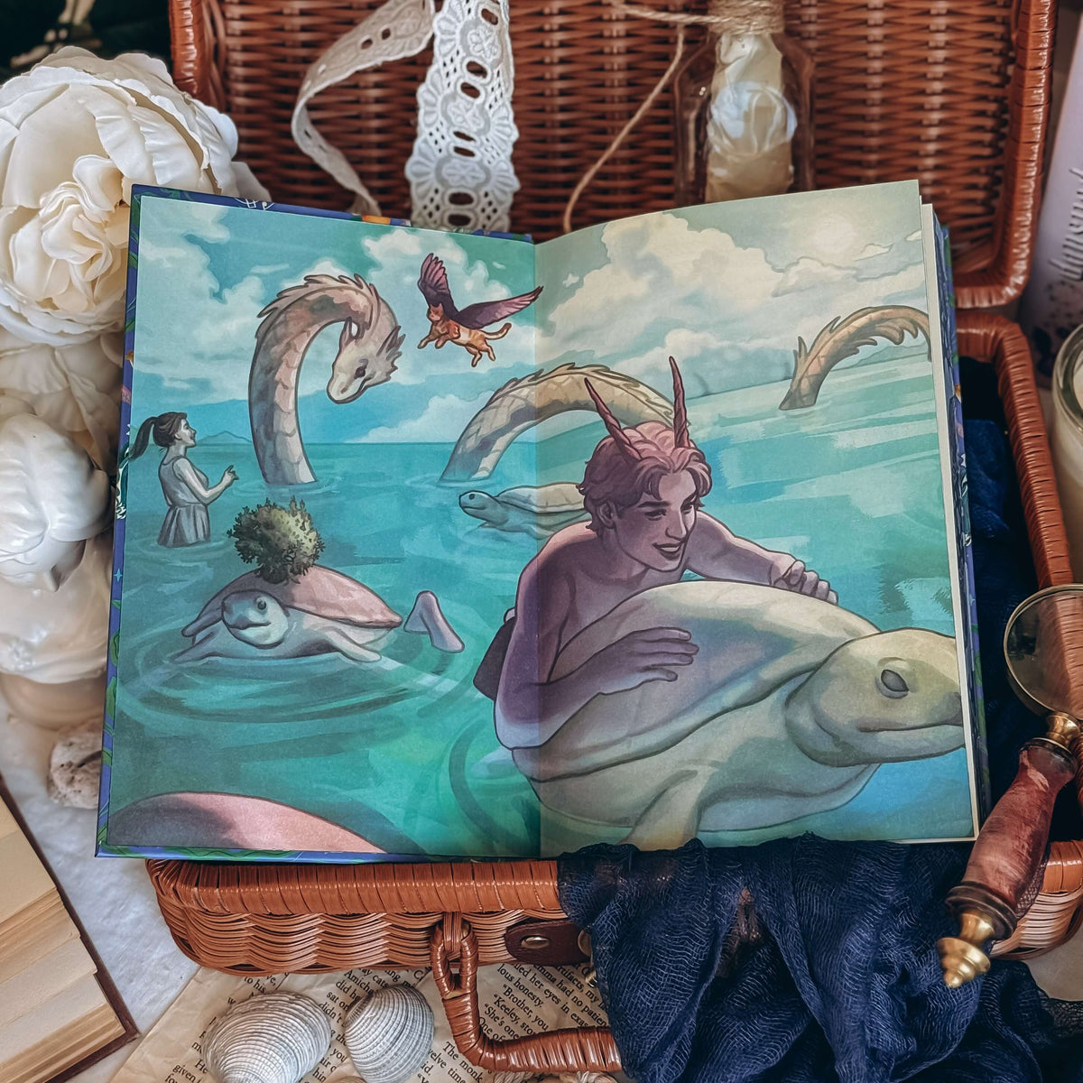 The front endpaper for the book Sea of Charms by Sarah Beth Durst shows an illustrated scene with a horned character riding a large sea turtle with a sea creature, a winged cat, another character and more sea turtles in the background. The book is styled in an open wicker basket with decorative flowers, lace, and dark blue fabric.