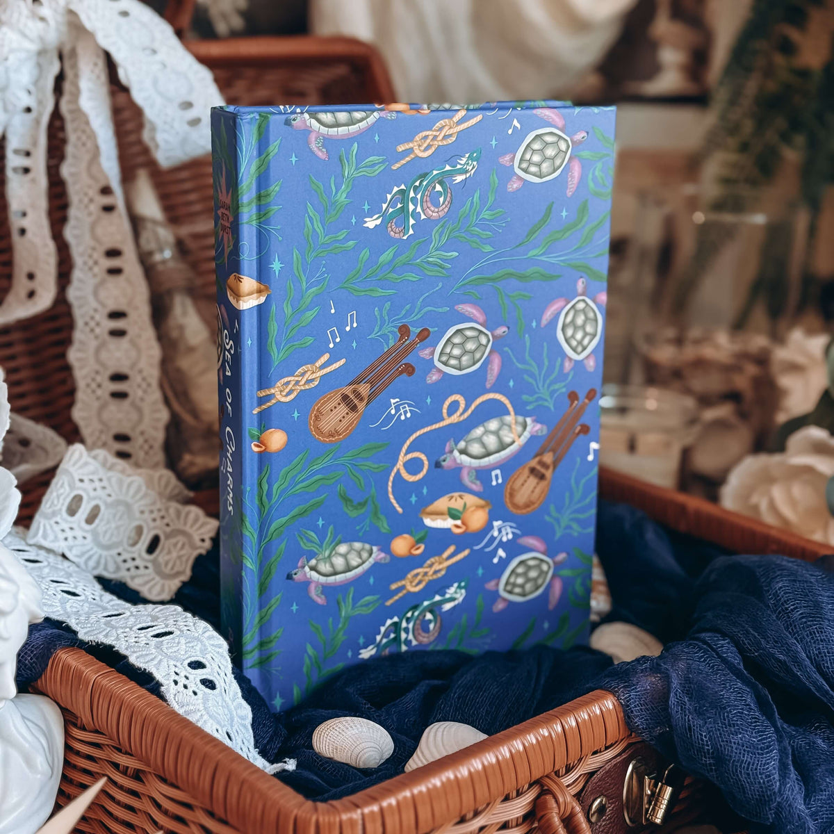 The hardcover for Sea of Charms by Sarah Beth Durst is feautres a whimsical ocean-themed illustration of sea turtles, kelp, and musical instruments, displayed inside a wicker basket with lace and fabric.