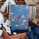 The hardcover for Sea of Charms by Sarah Beth Durst is feautres a whimsical ocean-themed illustration of sea turtles, kelp, and musical instruments, displayed inside a wicker basket with lace and fabric.