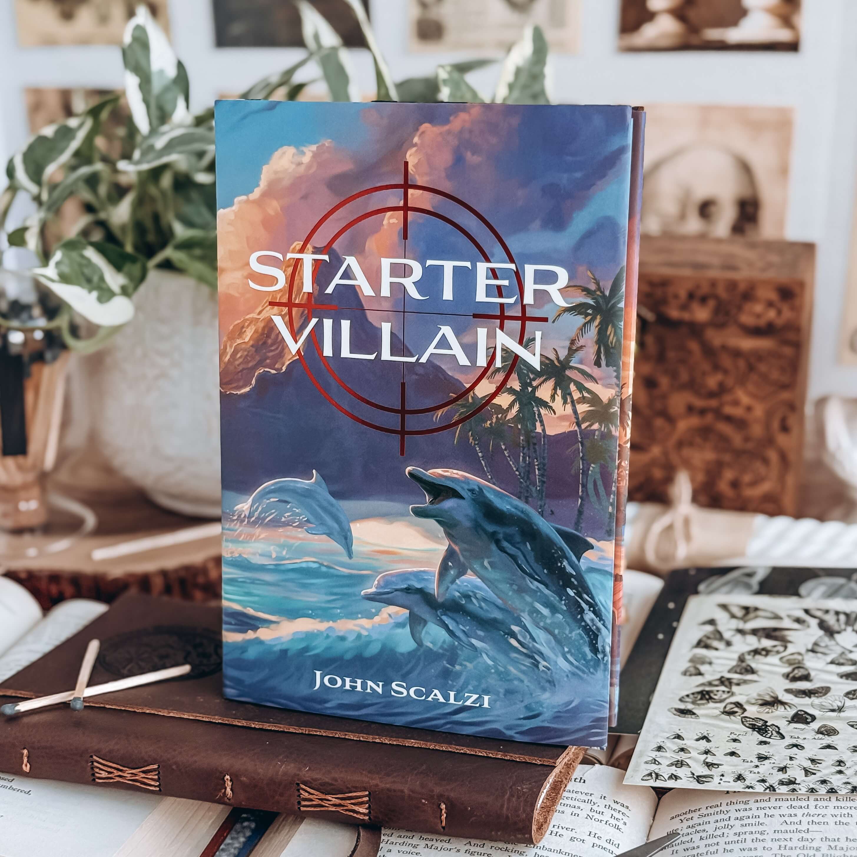 Starter Villain by John Scalzi. Cover shows dophins jumping out of the ocean with palm trees and mountains in the background. 