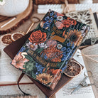 Hardcover of Starter Villain by John Scalzi. Decorated floral book features text reading "Dead? Suit it, motherfucker." Book is surrounded by decorative items. 