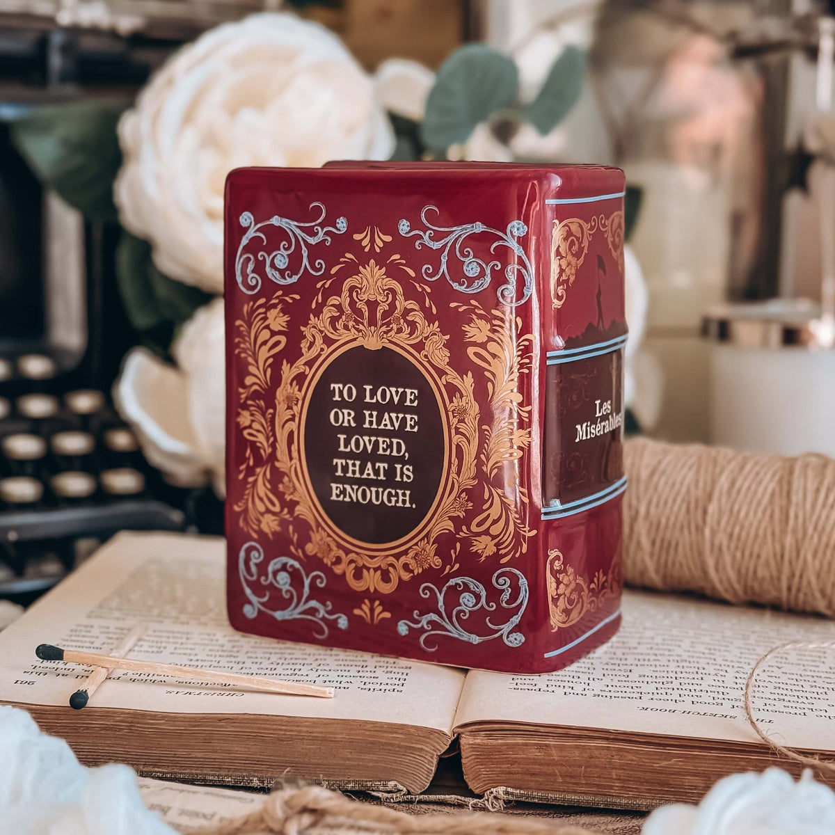 The back of a book-shaped vase modeled after a copy of Les Miserables sits on an open book with twine, white flowers, and a typewriter in the background.