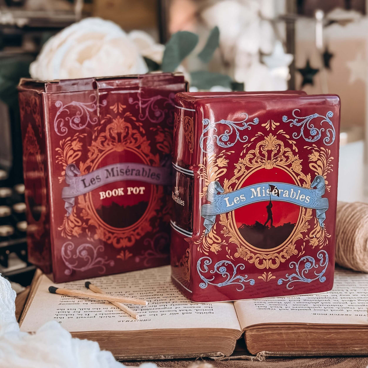 The front of a book-shaped vase modeled after a copy of Les Miserables and its matching cardboard box sits on an open book with twine, white flowers, and a typewriter in the background.