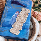 Sorcery of Thorns-inspired wooden bookmark with a purple tassle lies on top of a blue book.