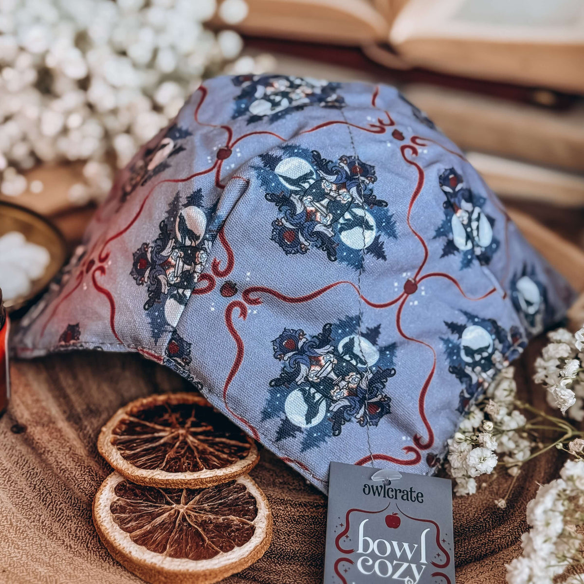A grey fabric bowl cozy with a repeating pattern featuring a Twilight-inspired crest, apples, and red ribbons lies upside down on a wooden base with dried orange slices and baby's breath flowers in surrounding.