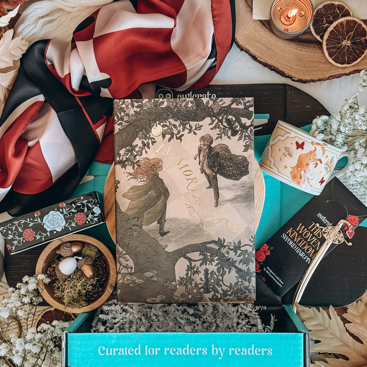 A styled flat-lay unboxing featuring a book titled Immortal Game by Allison Saft with an illustrated cover, surrounded by a chessboard-styled scarf, a ceramic trinket dish, a ceramic mug, and a sword hair pin, surrounded by various decorative items accessories.