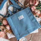 Ella Enchanted-inspired blue corduroy tote bag lies on white knit sweater surrounded by pink flowers.