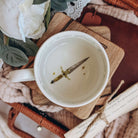 The topview of teacup with a dagger printed on the buttom of the cup sits on a stack of wooden coasters. The cup is surrounded by cream fabrics and white flowers.