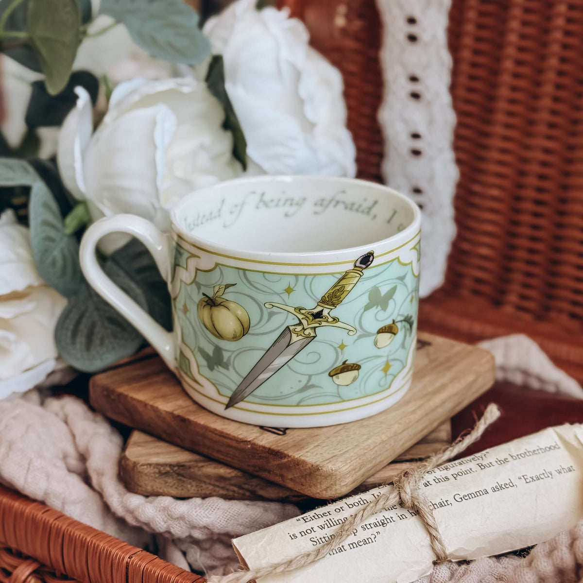 A teacup featuring a motif of daggers and acorns on a light teal background sits on a stack of wooden coasters. The cup is surrounded by cream fabrics and white flowers.