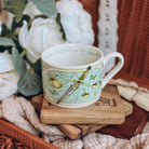 A teacup featuring a motif of daggers and acorns on a light teal background sits on a stack of wooden coasters. The cup is surrounded by cream fabrics and white flowers.