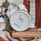 The bottom of the teacup is shown, with the Holly Black and OwlCrate logos printed. The mug sits on a stack of wooden coasters, surrounded by cream fabrics and white flowers.