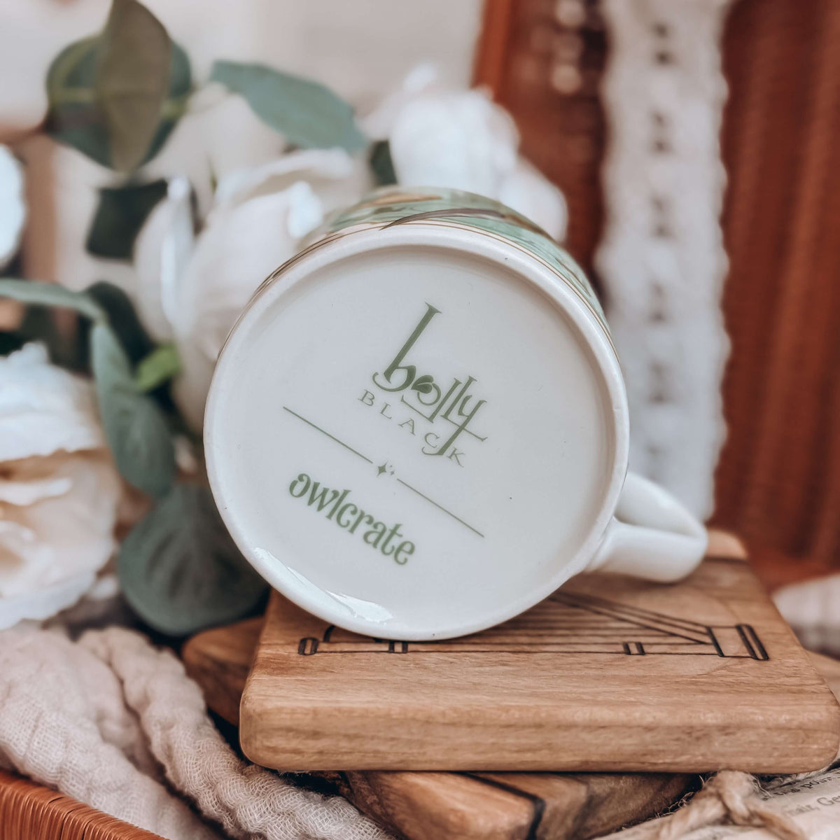 The bottom of the teacup is shown, with the Holly Black and OwlCrate logos printed. The mug sits on a stack of wooden coasters, surrounded by cream fabrics and white flowers.