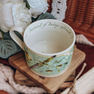 A closer view of the teacup showing a printed quote lining the inside of the mug sits on a stack of wooden coasters. The cup is surrounded by cream fabrics and white flowers.