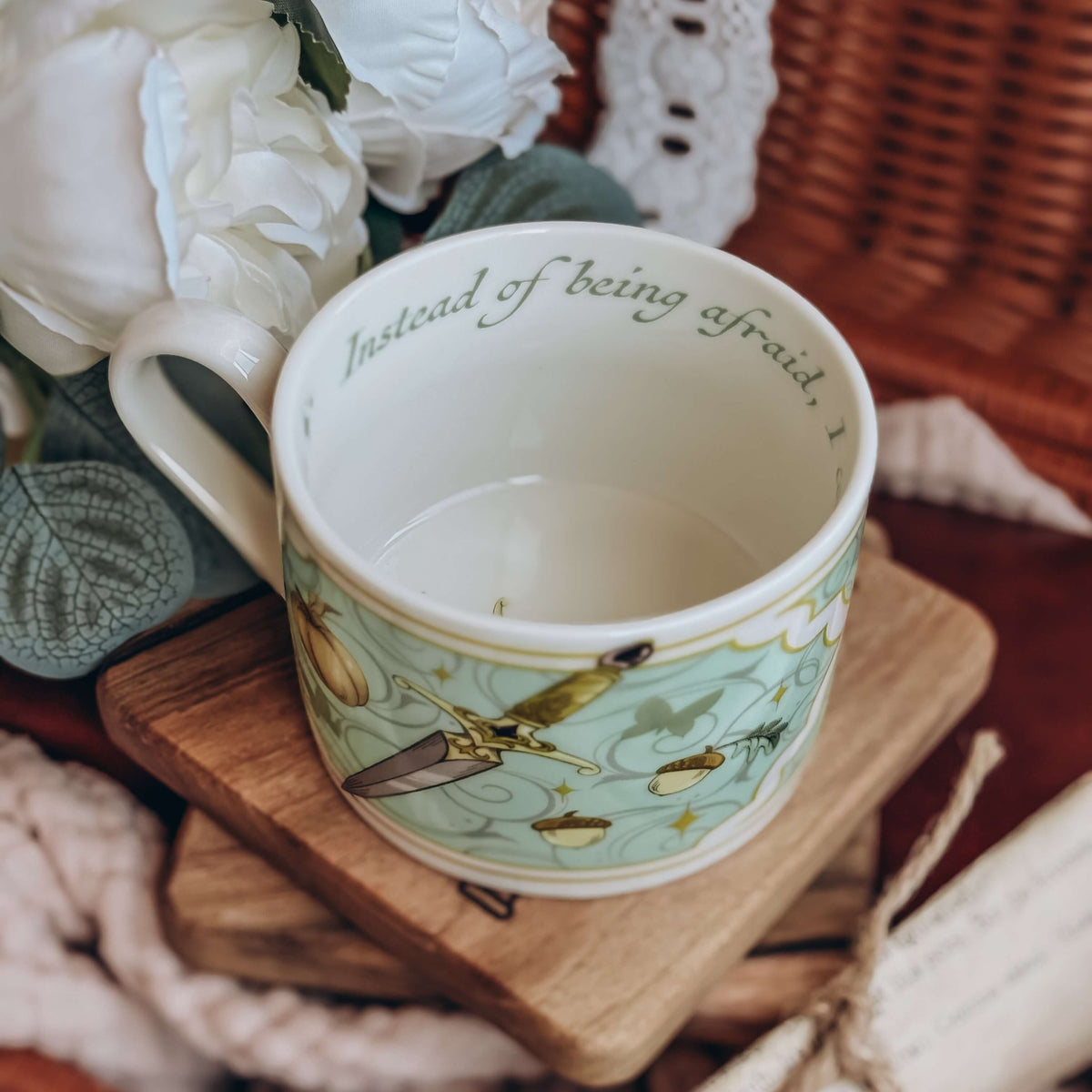 A closer view of the teacup showing a printed quote lining the inside of the mug sits on a stack of wooden coasters. The cup is surrounded by cream fabrics and white flowers.