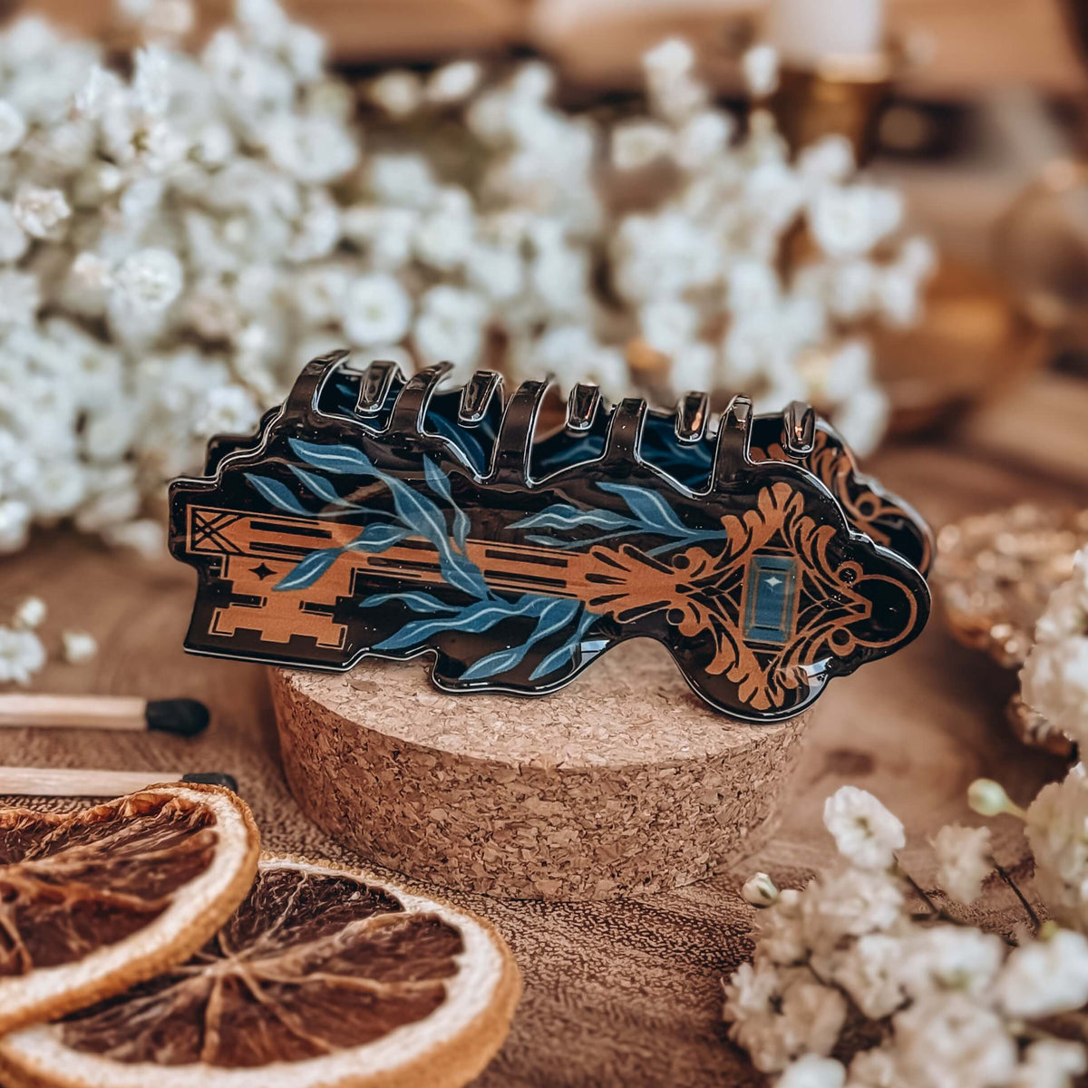A hair clip that resembles a skeleton key wrapped in kelp lies on a cork stand surrounded by dried orange slices, matchsticks, and baby's breath flowers.