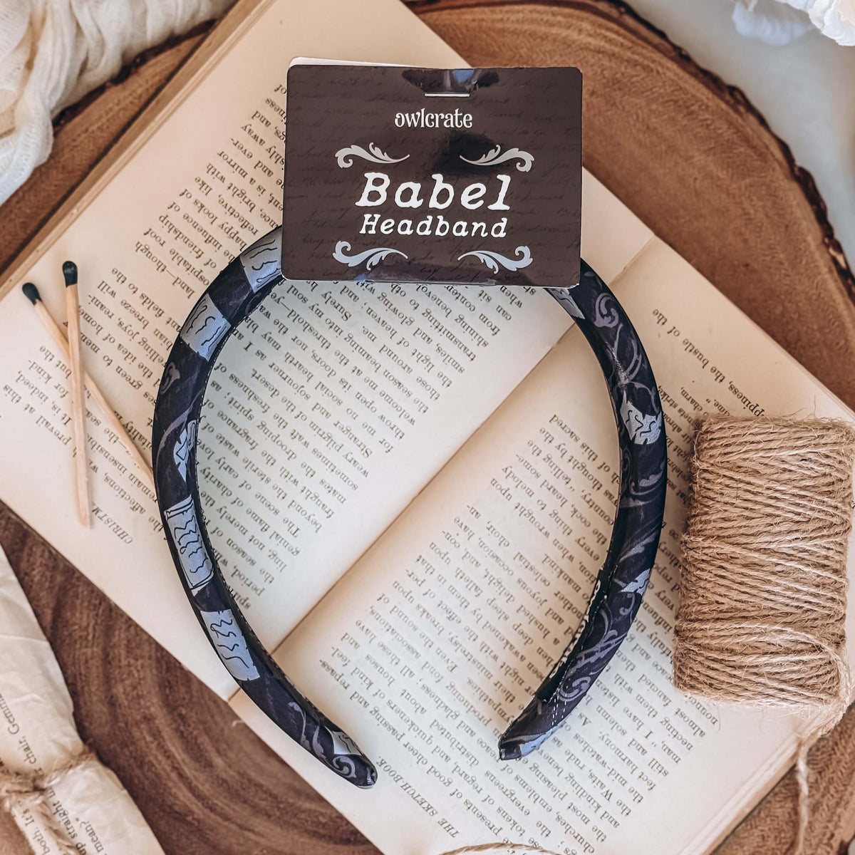 A black fabric headband with a pattern made of pages and decorative designs rests on an open book, surrounded by a roll of twine and matchsticks.