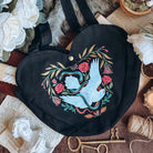 A black heart-shaped tote back featuring a a dove and serpent design is centered with keys, marble busts, books, twine, and white flowers surrounding.