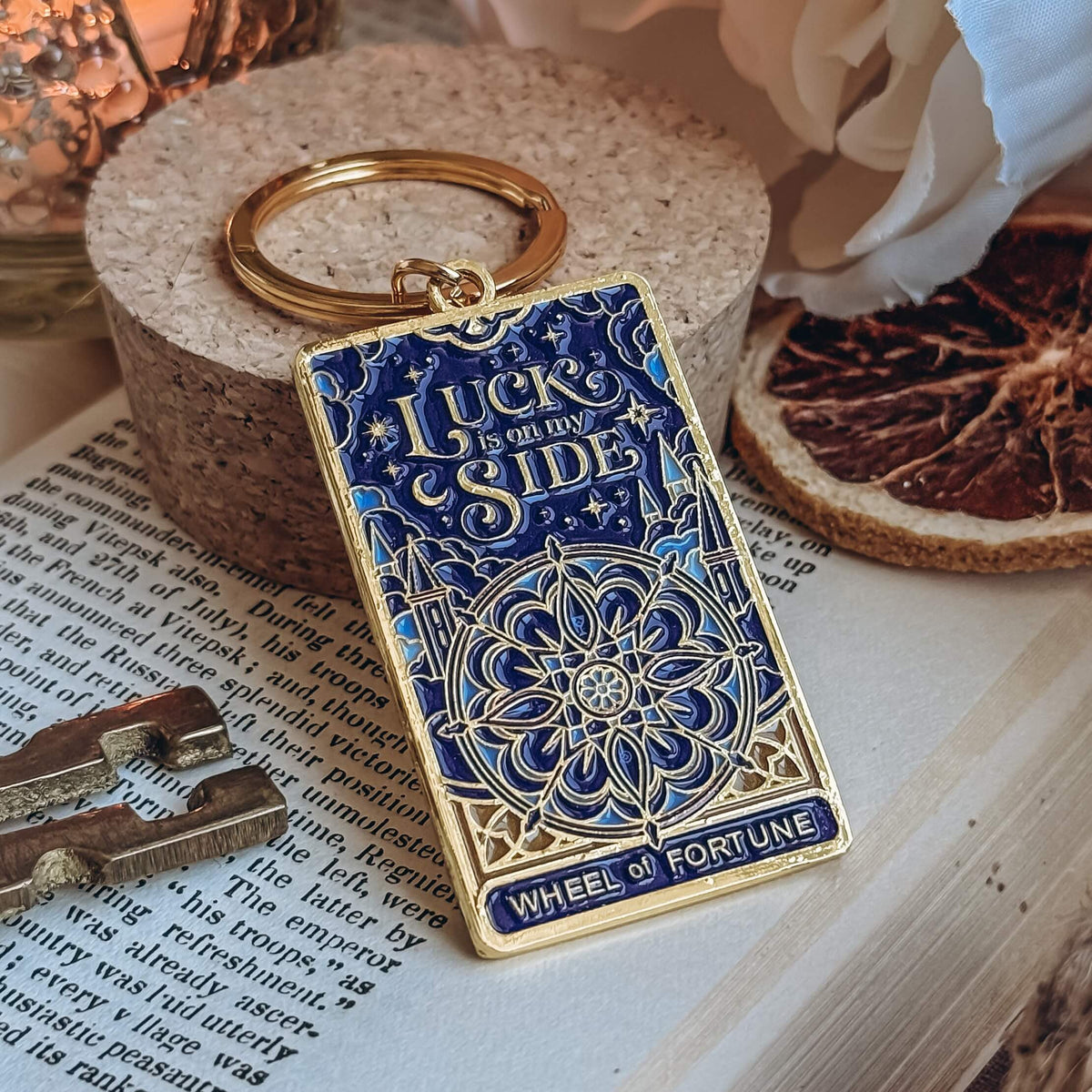 A blue and gold keychain designed to resemble the Wheel of Fortune tarot card rests on a cork stand on top of an open book. Surrounding the keychain is a skeleton key, a dried orange slice, and a white flower.