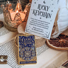 A blue and gold keychain designed to resemble the Wheel of Fortune tarot card rests on a cork stand next to a paper envelope for the Lucky Keychain. Both the keychain and the evelope are on top of an open book, surrounded by the keychain is a skeleton key, a dried orange slice, a white flower, and a lit candle in a glass votive.