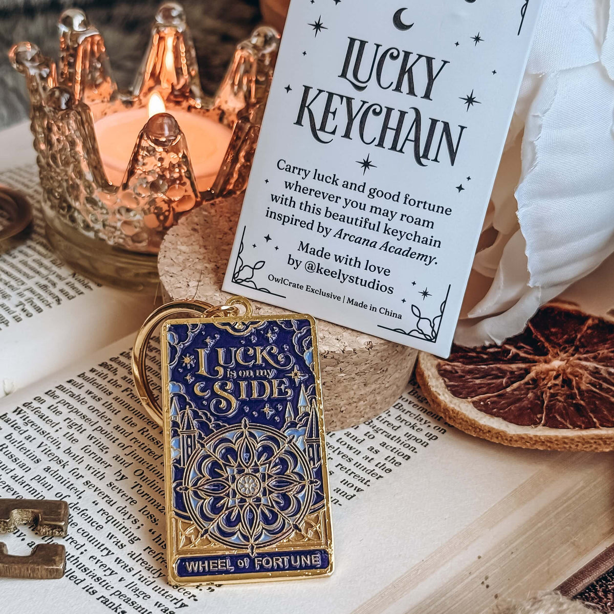 A blue and gold keychain designed to resemble the Wheel of Fortune tarot card rests on a cork stand next to a paper envelope for the Lucky Keychain. Both the keychain and the evelope are on top of an open book, surrounded by the keychain is a skeleton key, a dried orange slice, a white flower, and a lit candle in a glass votive.