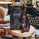 A carton of Fantasy Artist Markers and a pile of five of those markers rest on a wooden stand. An open book, a typewriter, and candles are visible in the background.