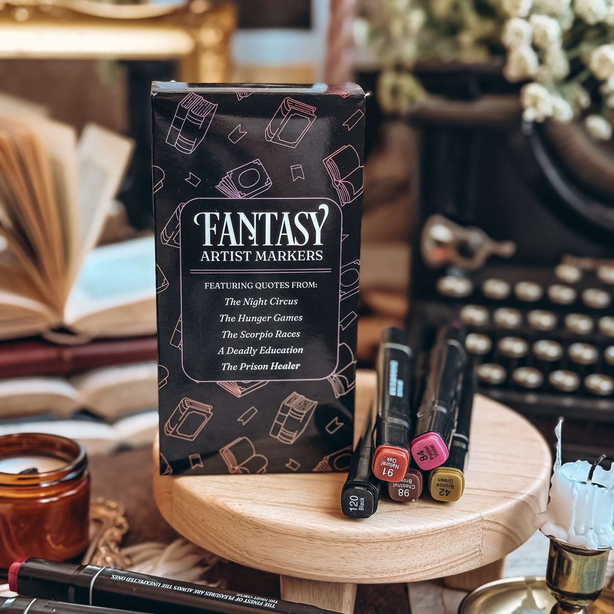 A carton of Fantasy Artist Markers and a pile of five of those markers rest on a wooden stand. An open book, a typewriter, and candles are visible in the background.