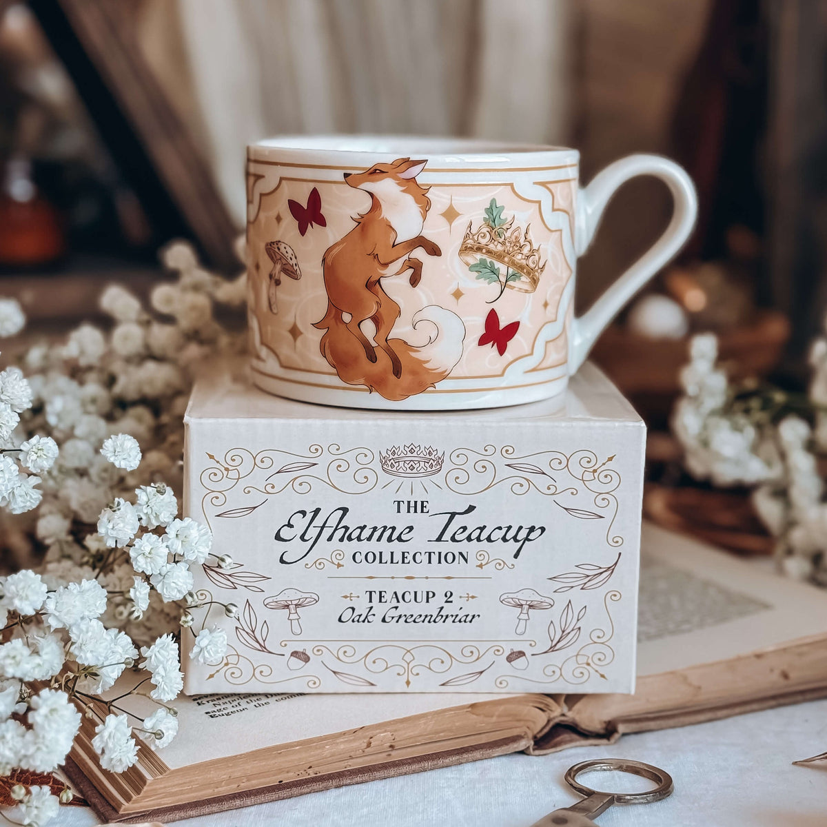 A cermaic teacup sits on top of a decorative product box, styled on an open book surrounded by baby's breath flowers. The teacup features a red fox on a pink framed background with gold detailing, circled by mushrooms, a gold crown, and red butterflies.