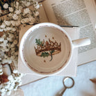 The topview of a white ceramic teacup sits on a white box, styled on an open book next to baby's breath flowers. The insde of the teacup shows an illustrated gold crown and a small, green oak leaf branch. 