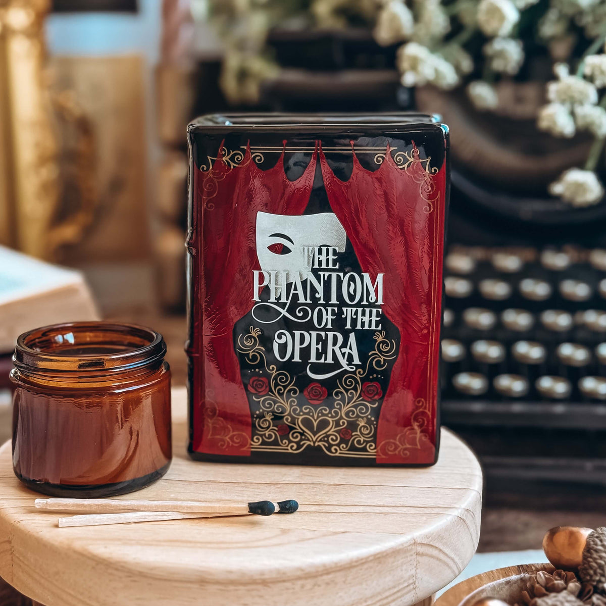 The front of a Phantom of the Opera-inspired book-shaped vase sits on a wooden stand next to a glass candle votive and two matchsticks. Book pot shows a phantom half-mask with the Phantom of the Opera title featured.