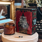 The back of a Phantom of the Opera-inspired book-shaped vase sits on a wooden stand next to a glass candle votive and two matchsticks. Book pot features a quote from the stage musical, "Let your soul take you where you want to be."