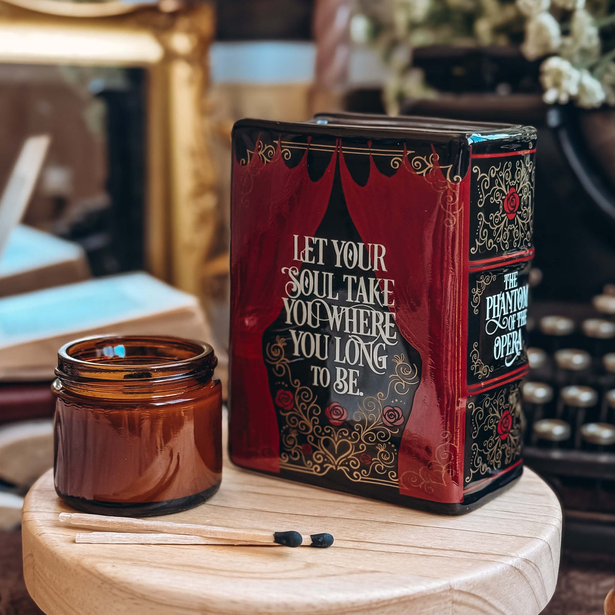 The back of a Phantom of the Opera-inspired book-shaped vase sits on a wooden stand next to a glass candle votive and two matchsticks. Book pot features a quote from the stage musical, "Let your soul take you where you want to be."