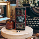 The spine of a Phantom of the Opera-inspired book-shaped vase sits on a wooden stand next to a glass candle votive and two matchsticks.