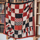 A square black, white, and red chessboard-inspired scarf is featured, draped on a brown bookshelf. 