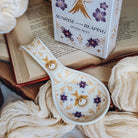 A spoon rest designed with a gold mocking jay, purple flowers, and gold art deco-inspired designs lies on a linen scarf next to a matching cardboard box on an open book.