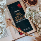 A gold decorative sword-shaped hair pin sits on a black branded envelope featruing the text 'OwlCrate, This Woven Kingdom sword hair pin.' Both rest on an open book surrounded by baby's breath flowers and dried orange slices. 