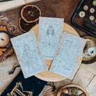 Three gold-foiled tarot cards, depicting the Main Character, Librarian, and Enemies to Lovers, lay on a wooden stand surrounded by candles, a typewriter, and papers.