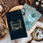 An OwlCrate bookish tarot velvet bag and three teal-coloured cards rest on a wooden stand surrounded by candles, succulents, and papers.