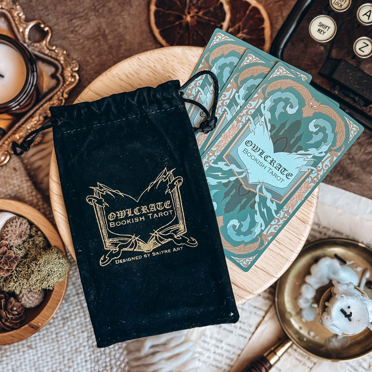 An OwlCrate bookish tarot velvet bag and three teal-coloured cards rest on a wooden stand surrounded by candles, succulents, and papers.