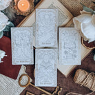 Four bookish-themed tarot cards lay across an open book, surrounded by keys, twine, a leather-bound book, a lit candle, and white flowers. The cards show tarot-inspired illustrated depictions of The Slow Burn, The Quest, The Prologue, and The Mood Reader.