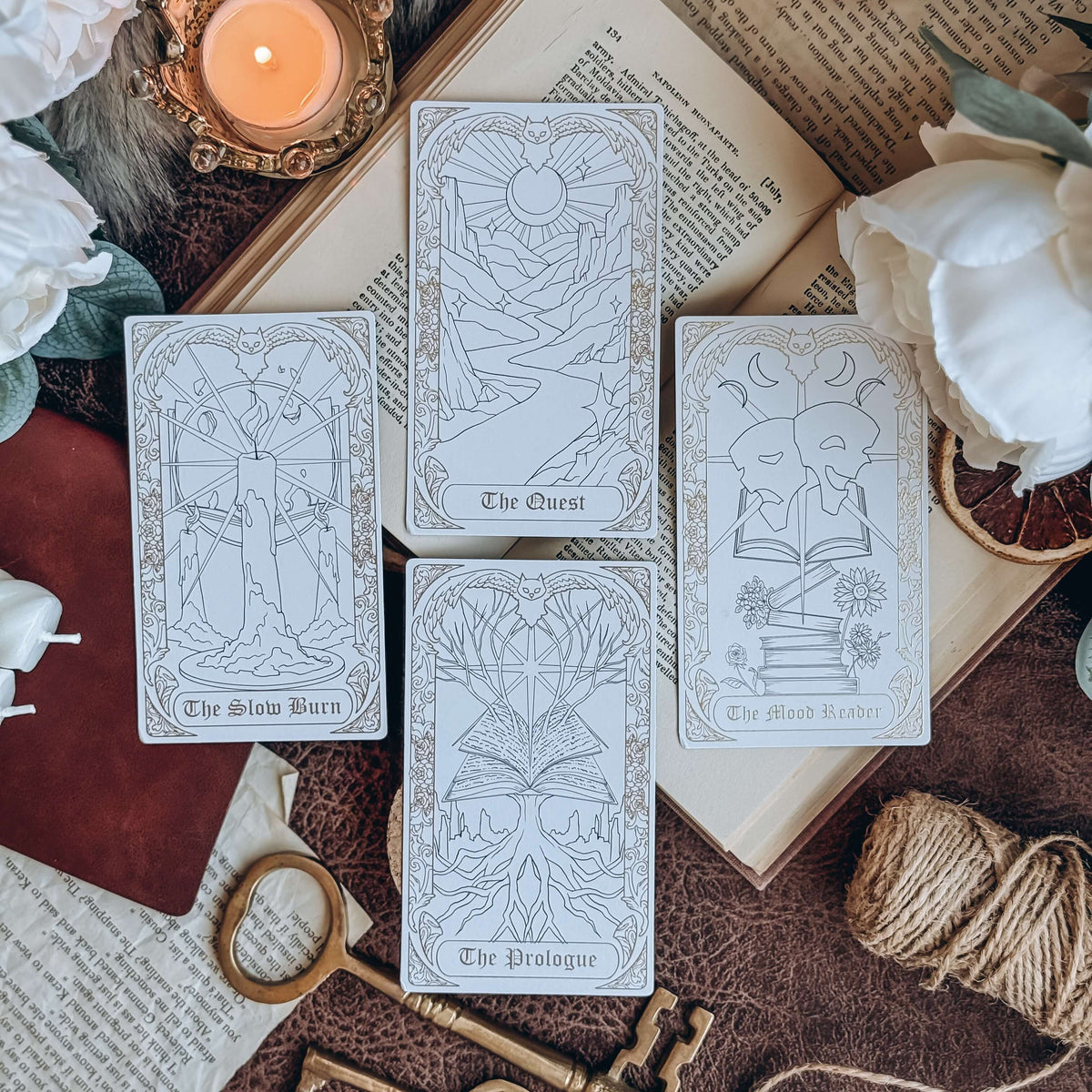 Four bookish-themed tarot cards lay across an open book, surrounded by keys, twine, a leather-bound book, a lit candle, and white flowers. The cards show tarot-inspired illustrated depictions of The Slow Burn, The Quest, The Prologue, and The Mood Reader.