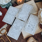 Four bookish-themed tarot cards lay diagonally across an open book, surrounded by keys, twine, a leather-bound book, a lit candle, and white flowers. The cards show tarot-inspired illustrated depictions of The Slow Burn, The Quest, The Prologue, and The Mood Reader.